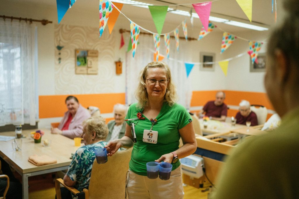 A caregiver serving drinks to elderly residents in a cheerful nursing home setting.